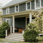 An older home with green-grey siding and off-white accents is a great example of a fixer-upper that has increased in value