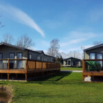 What a community might look like when buying a mobile home. A few green mobile homes with black trim sit on stained decks in a grassy field near a pond on a clear blue day.