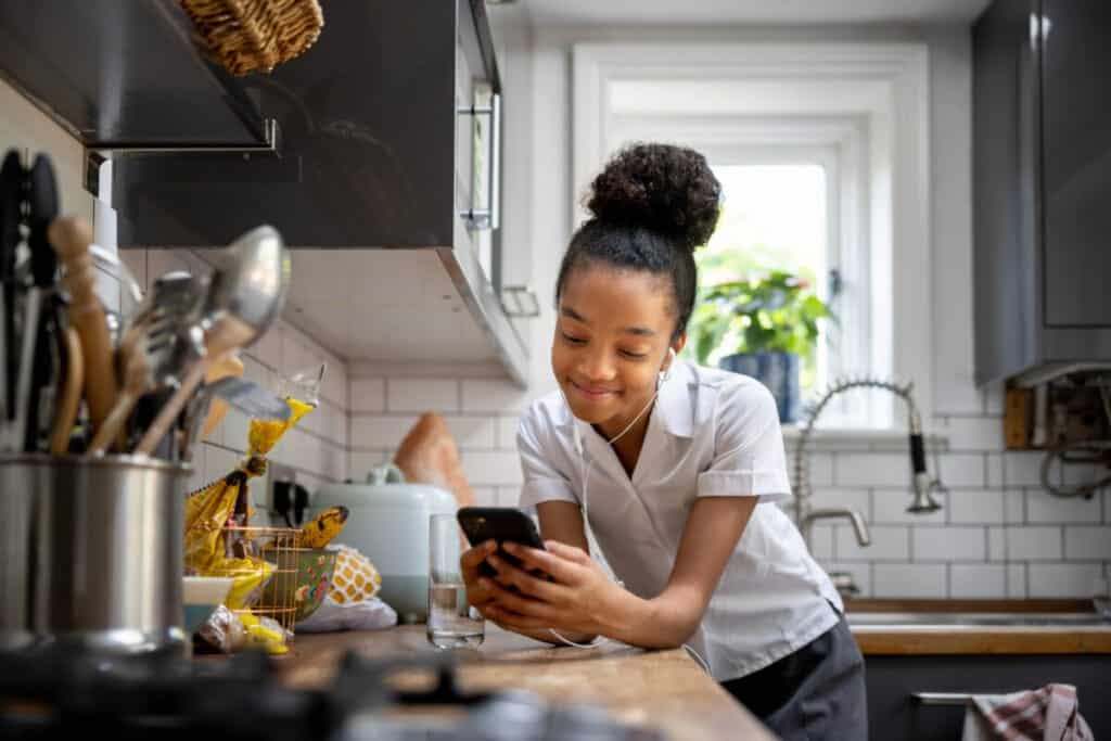 Teenage girl at home texting on her cell phone while drinking a glass of water in the kitchen