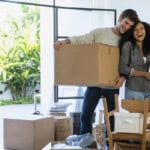 Happy couple carrying cardboard box and pot plant in new house