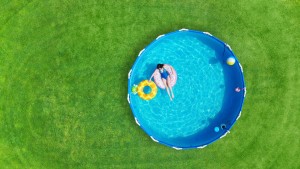 Aerial. Girl swims in a metal frame pool with inflatable toys. Frame pool stand on a green grass lawn. Top view. Copy space.
