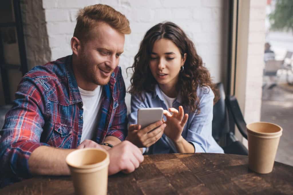 House hunting is part of the process when buying a house with a friend. Two friends sit at a coffee shop and look at listings on a phone.