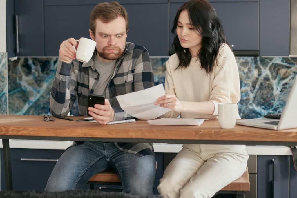Man looking concerned while holding a white mug and phone, sitting at a wooden table across from a woman reviewing papers, with a laptop nearby in a modern kitchen. Appraisal came in low.