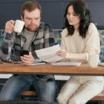 Man looking concerned while holding a white mug and phone, sitting at a wooden table across from a woman reviewing papers, with a laptop nearby in a modern kitchen. Appraisal came in low.