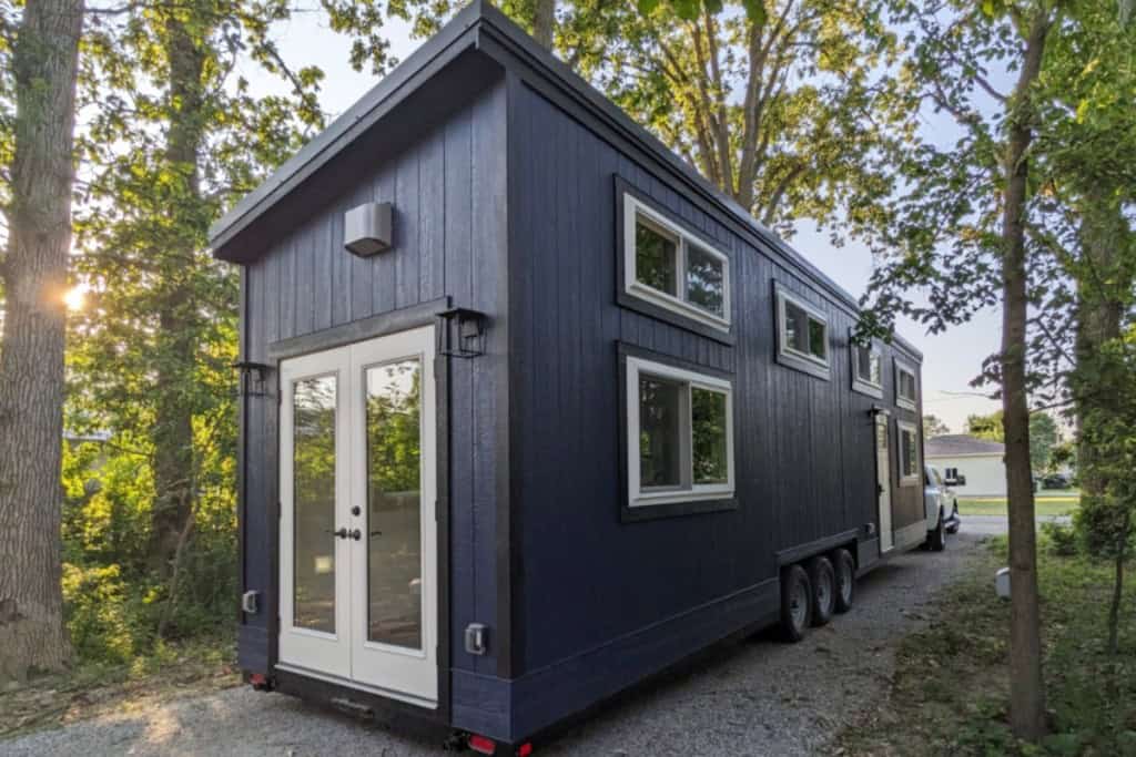 A dark blue granny pod, a type of tiny home, is parked on a gravel path surrounded by trees. The granny pod has a sloped roof, white-framed windows of varying sizes, and double white doors at the end.