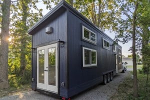A dark blue granny pod, a type of tiny home, is parked on a gravel path surrounded by trees. The granny pod has a sloped roof, white-framed windows of varying sizes, and double white doors at the end.