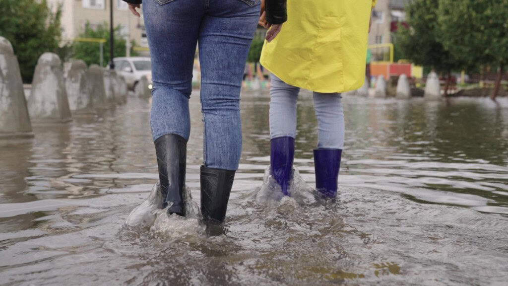 Two people with high waterproof boots walk through a flooded street in a neighborhood in a flood zone.
