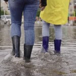 Two people with high waterproof boots walk through a flooded street in a neighborhood in a flood zone.