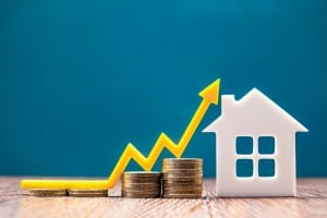 A white, cutout-style house sits on a wooden surface next to stacks of gold coins that gradually increase in height, representing growing equity or value. A bright yellow arrow dramatically curves upwards from the lowest stack of coins, pointing towards the top right, symbolizing an increase. The background is a solid, deep teal color. This image visually represents the concept of increasing home equity and financial growth, which is often a consideration when asking "Do you need a down payment to refinance?"