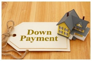 A miniature yellow and grey house sits next to a price tag-shaped label tied with twine. The label clearly reads "Down Payment" in a bold, golden font on a light background. This image highlights the concept of a minimum down payment for a house for a first-time buyer, all resting on a warm, wooden surface.
