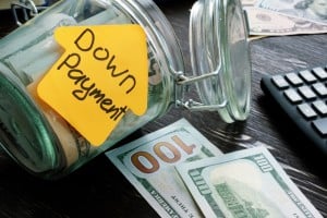 A glass jar, labeled with a yellow sticky note shaped like a house and the words "Down Payment," is tipped on its side, with money spilling out. Several $100 bills are visible outside the jar, alongside a calculator and more scattered currency in the background, all on a dark wooden surface. This image prompts the question, "Is a Large Down Payment on a House Always Best?"