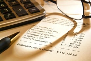 A close-up shot of a document detailing estimated cash at closing, with line items for various fees like state transfer tax, county recording fees, and broker's commission. The total estimated cash at closing is $182,530.00. A calculator and a pair of eyeglasses are blurred in the background, suggesting a financial or legal context.