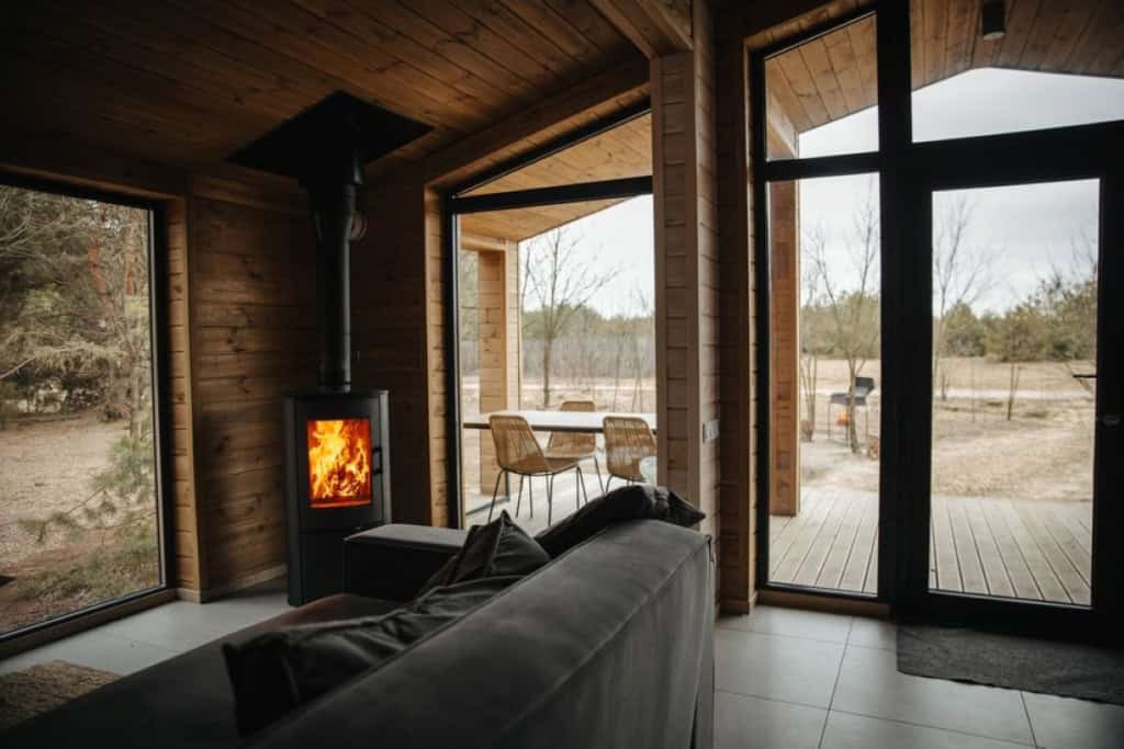 Modern wood-burning stove with fire inside a cozy cabin living room featuring large windows and natural wood walls, overlooking outdoor patio and trees.