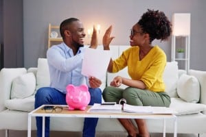 A happy couple high-fives while looking at paperwork and a pink piggy bank on a coffee table. The image represents the joy of successfully navigating a financial decision, such as making a winning "highest and best offer" on a home.