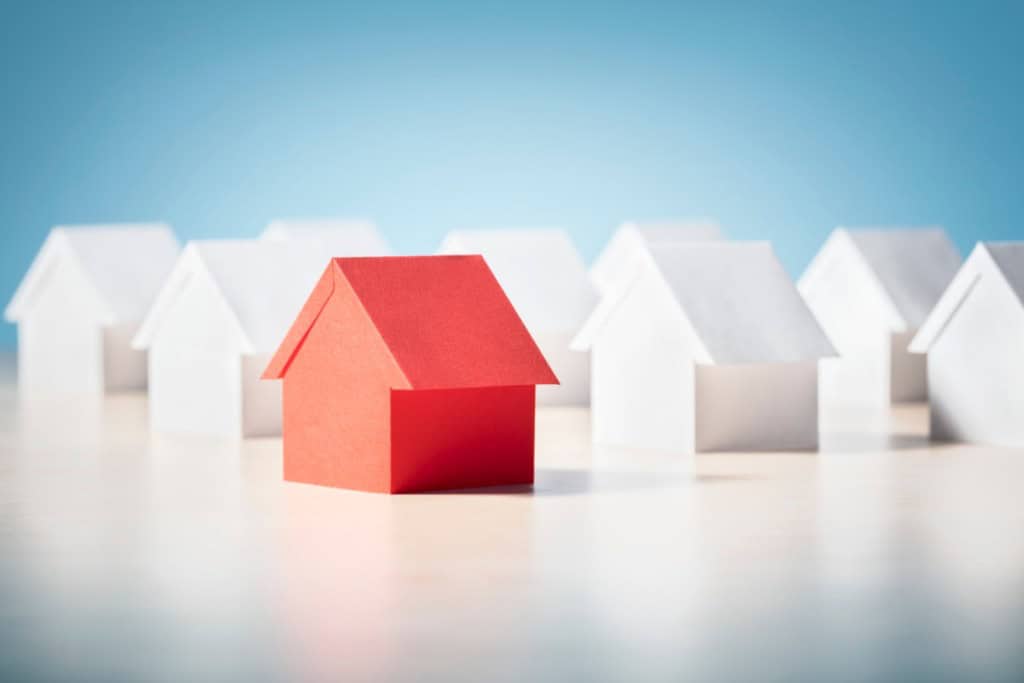 A single red paper house stands out from a row of white paper houses, symbolizing the challenge of winning a bid in a competitive market. The image represents the theme of "how to buy a home in a seller's market" by visually highlighting a unique, standout option among many similar choices.