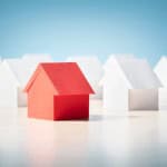 A single red paper house stands out from a row of white paper houses, symbolizing the challenge of winning a bid in a competitive market. The image represents the theme of "how to buy a home in a seller's market" by visually highlighting a unique, standout option among many similar choices.