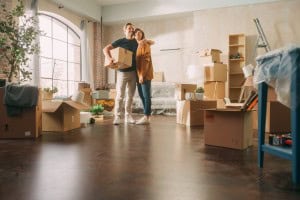 A smiling couple embraces in their new home, surrounded by moving boxes, a ladder, and houseplants. They are standing on a polished wood floor in a sunlit room with an arched window, capturing a happy moment during the process of moving in.non contingent offer