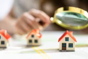 A person holds a magnifying glass over a miniature house on a map, with two other small houses nearby. The image symbolizes a real estate agent identifying and examining **expired listing leads for agents**.
