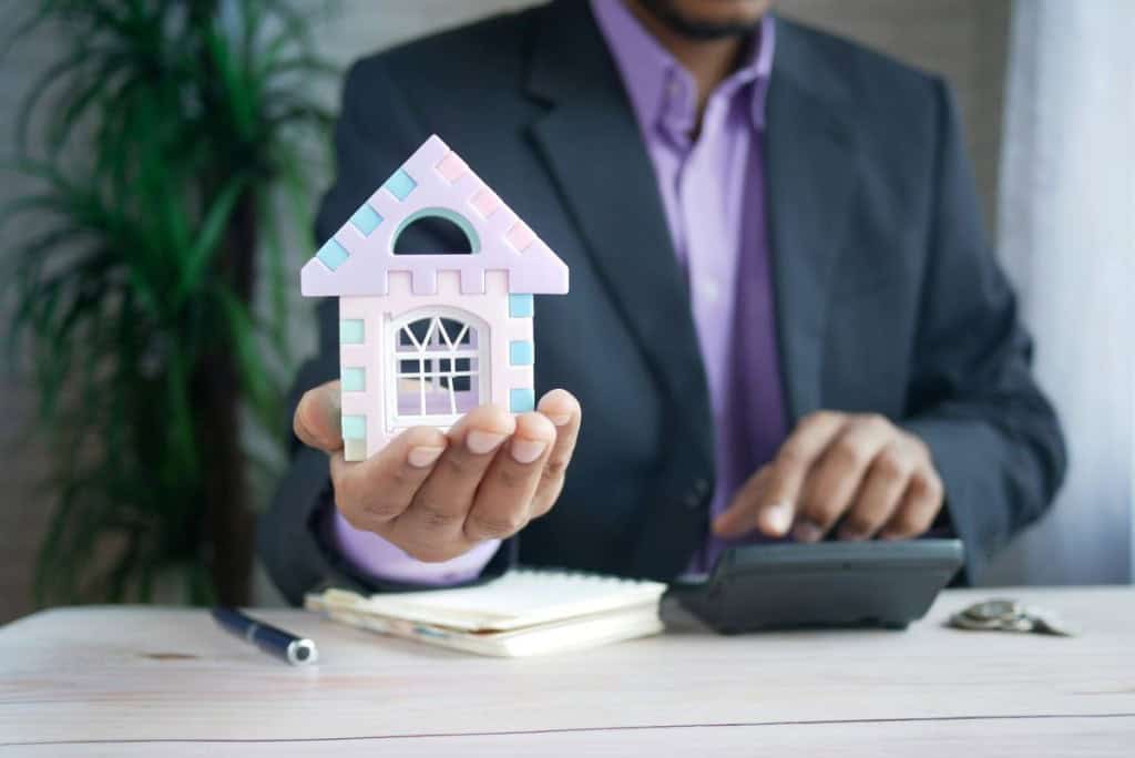 A man in a suit holding a toy house and typing on a calculator to determine if capital gains taxes need to be paid on a house sale