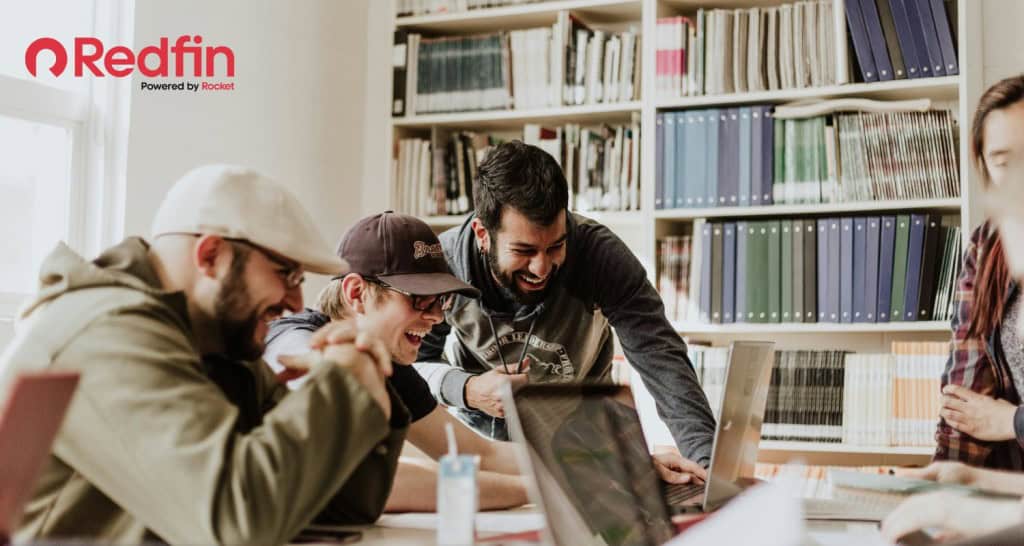 Use Redfin to learn how to rent an apartment as an international student. A group of students are gathered around a laptop in a study hall. They are laughing together.