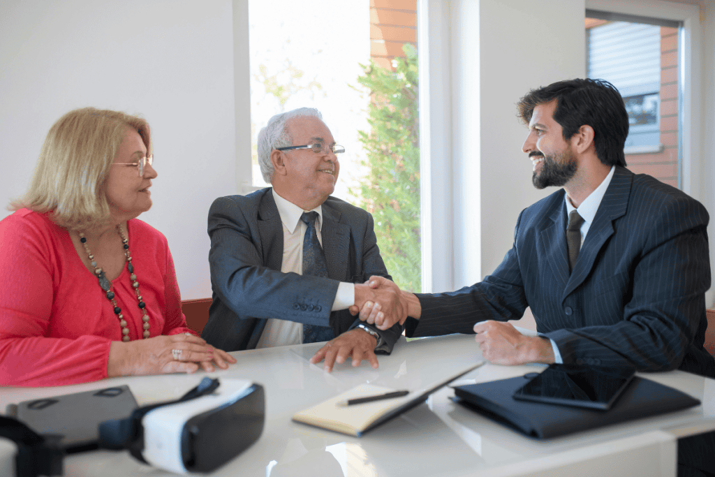 A well-dressed real estate agent sits at a bright office table, smiling as he shakes hands with an older man in a suit, while an older woman sits beside him watching the exchange. Sunlight streams through large windows behind them. Papers, a tablet, a notebook, and a virtual reality headset rest on the table, suggesting a modern home buying discussion. The scene reflects trust and professionalism in the process of How to Protect Your Client During Underwriting.